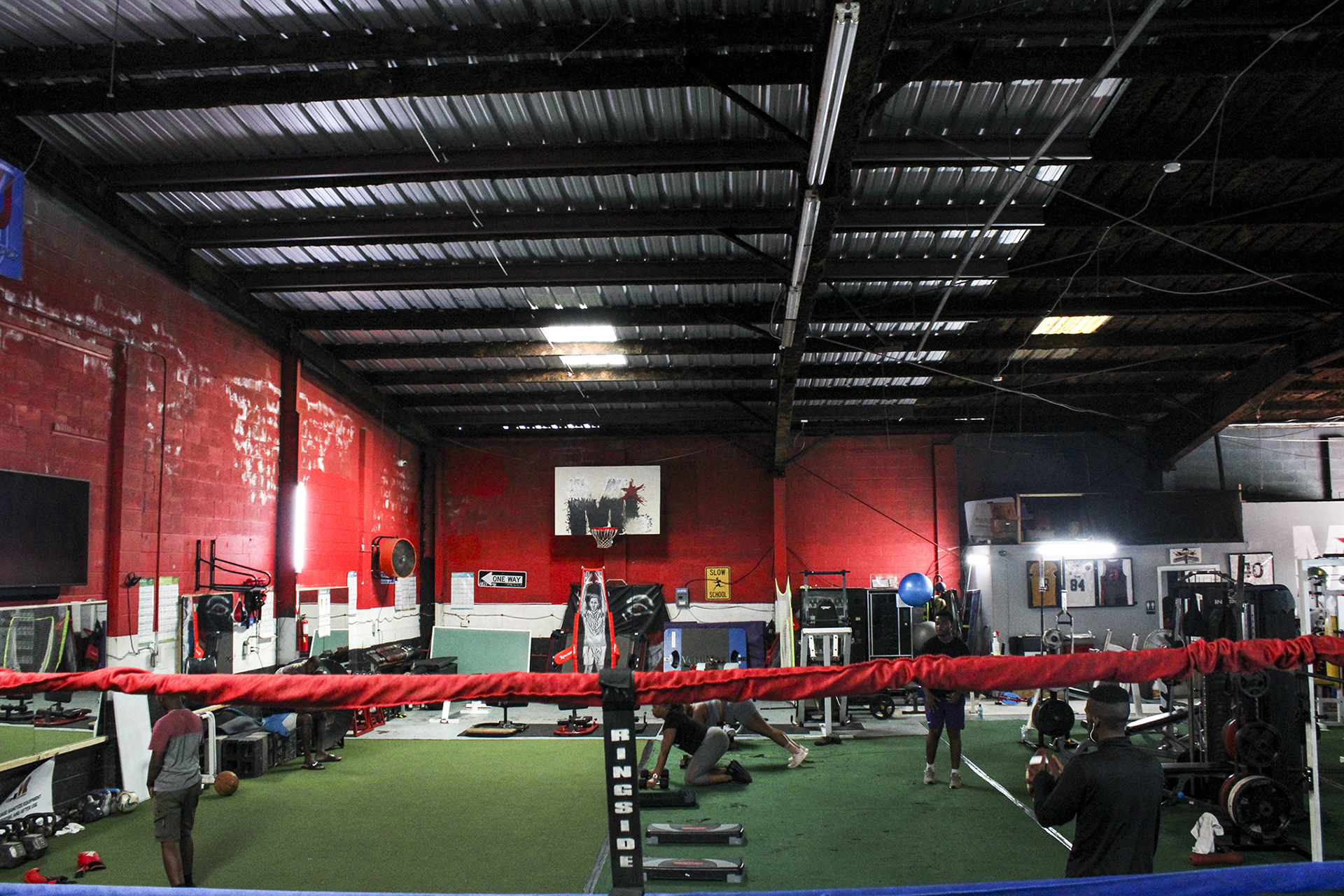 Red and blue ropes from inside a boxing ring in MT Athletics in Uptown New Orleans. The walls are red, the ceilings are black, the floor is green, there is a basketball hoop hanging up and there are people exercising.