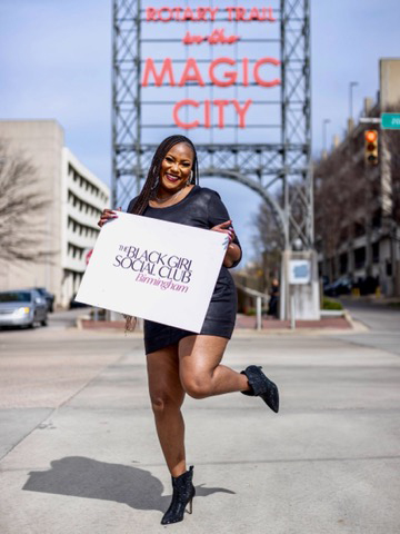 A Black woman is holding a cardboard sign that reads, “The Black Girl Social Club Birmingham.” She is standing in front of a walkway entrance labeled “Rotary Trail in the Magic City.”