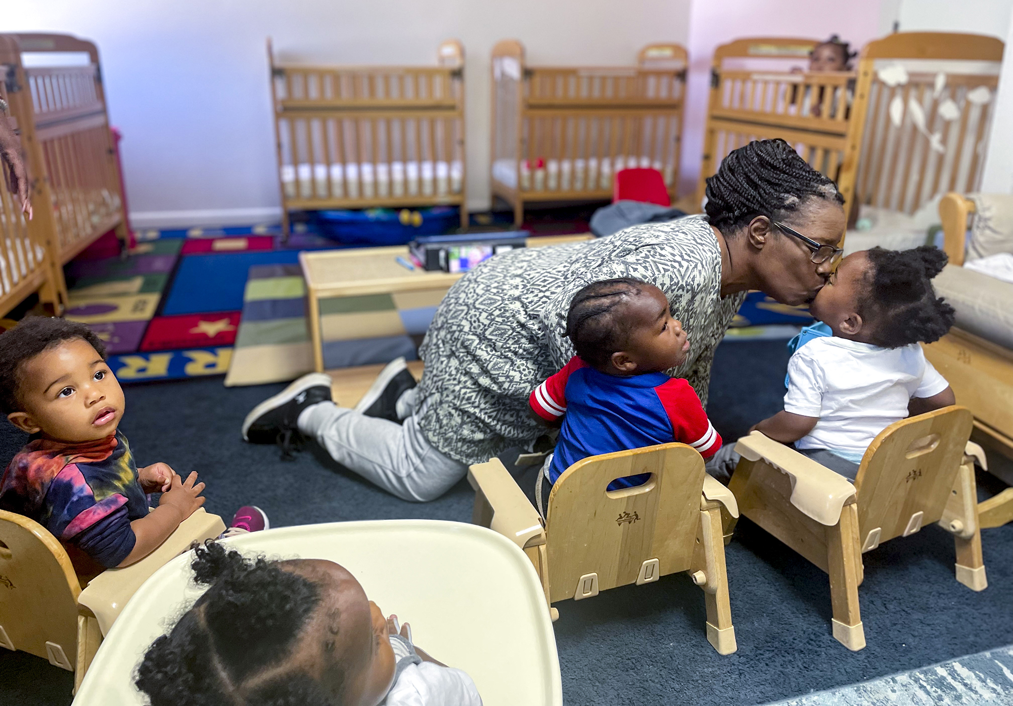 Deborah Holmes wears glasses and leans over to kiss a toddler who’s seated in a wooden chair. Other children are seated. 